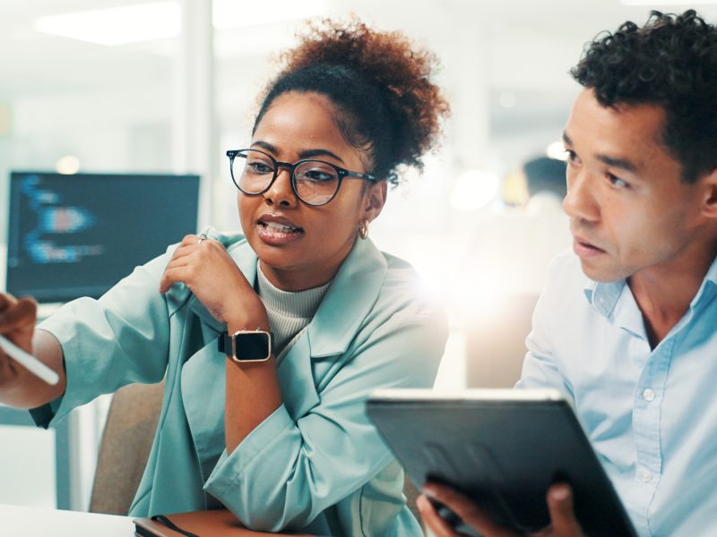 Man holding a tablet sitting next to a woman who is pointing at a desktop monitor