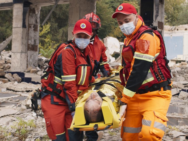 Three members of an emergency service team carry a person on a stretcher away from an area of fallen masonry